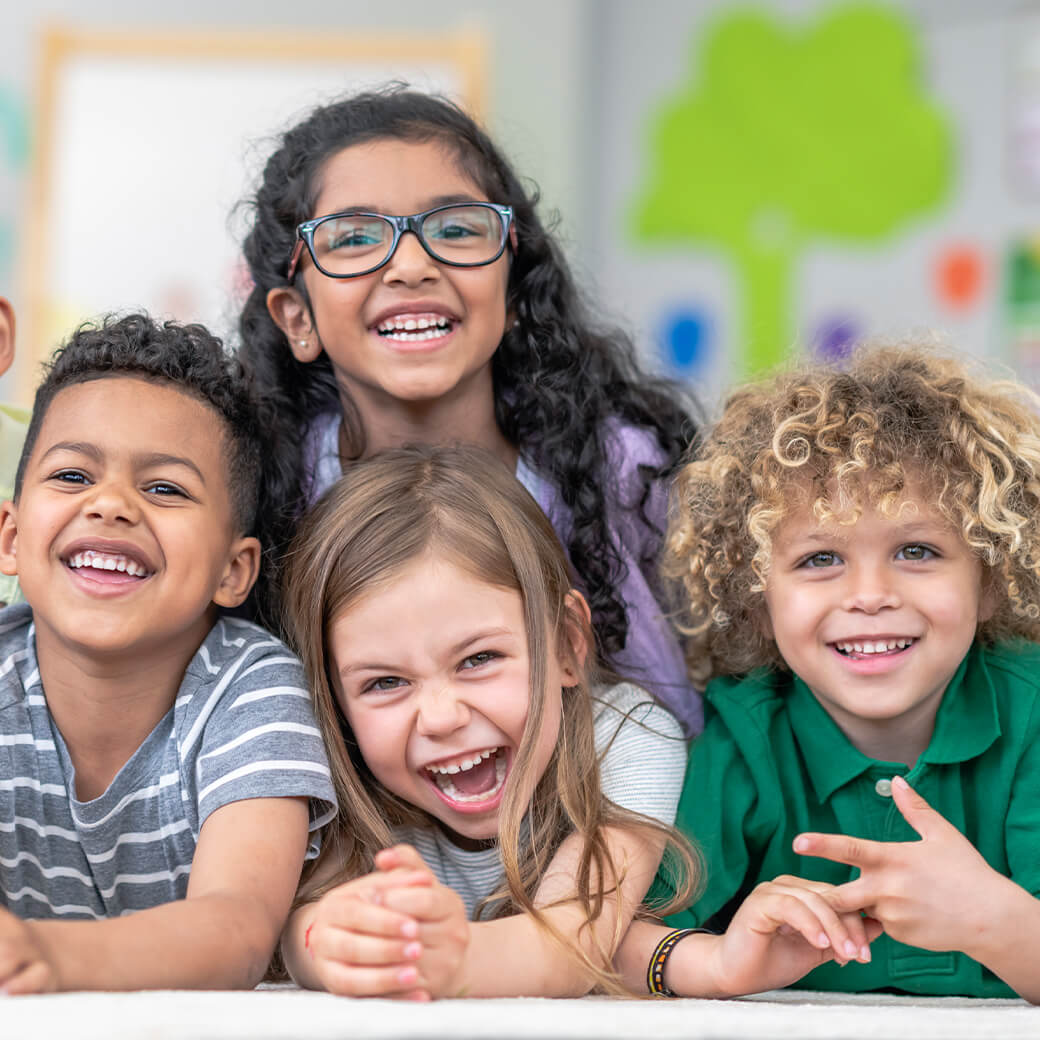 Group of smiling and laughing children laying in a pile on the ground in a classroom.