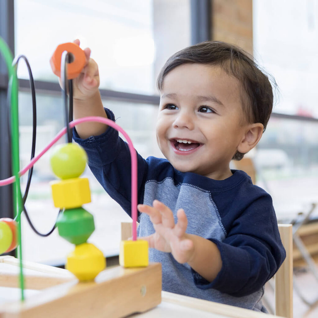 Toddler boy sits at a table, reaching up cheerfully to play with a toy bead maze.